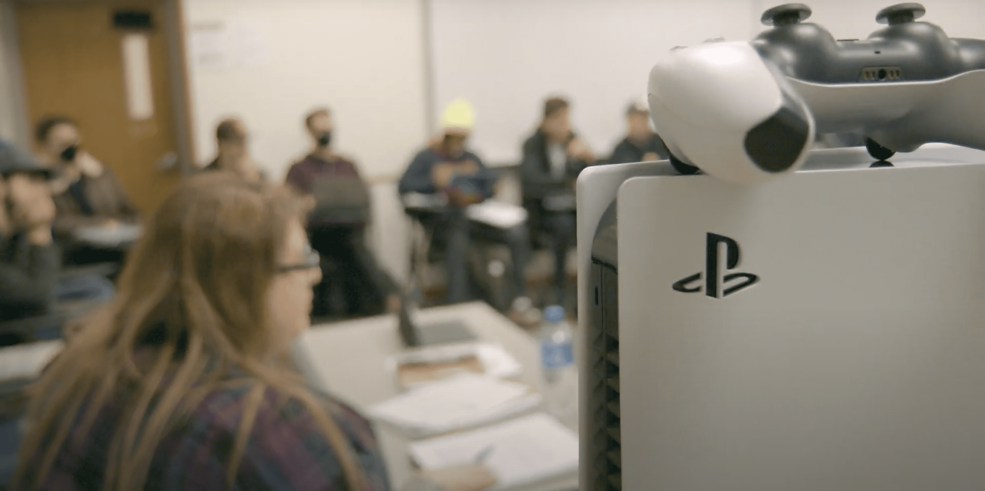 Class A close up shot of a PS5, with the college professor and her students in the background, out of focus.