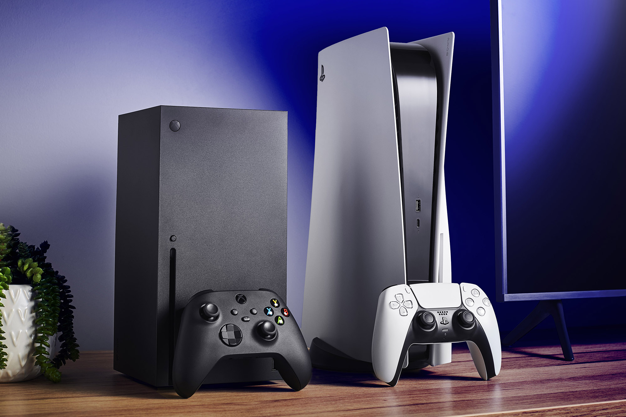 Xbox Series X and PS5 consoles with controllers on a wooden table.