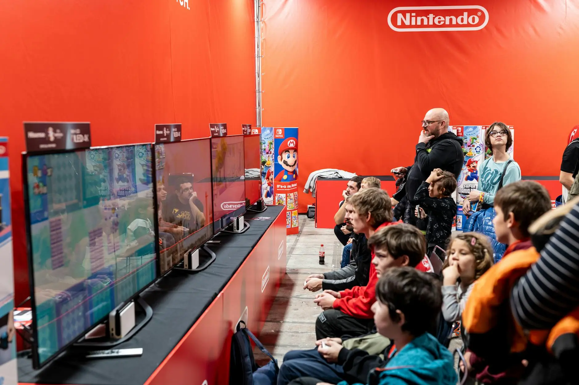 Crowd watching Nintendo games on multiple screens at an event with a red background.