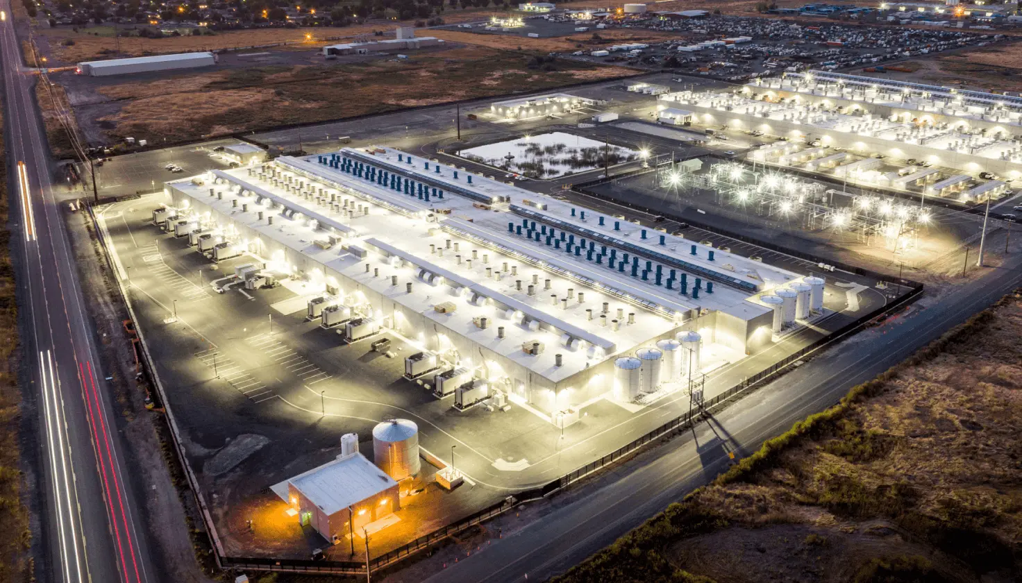 Aerial night view of a large industrial data center complex with rows of white cooling units and bright lighting
