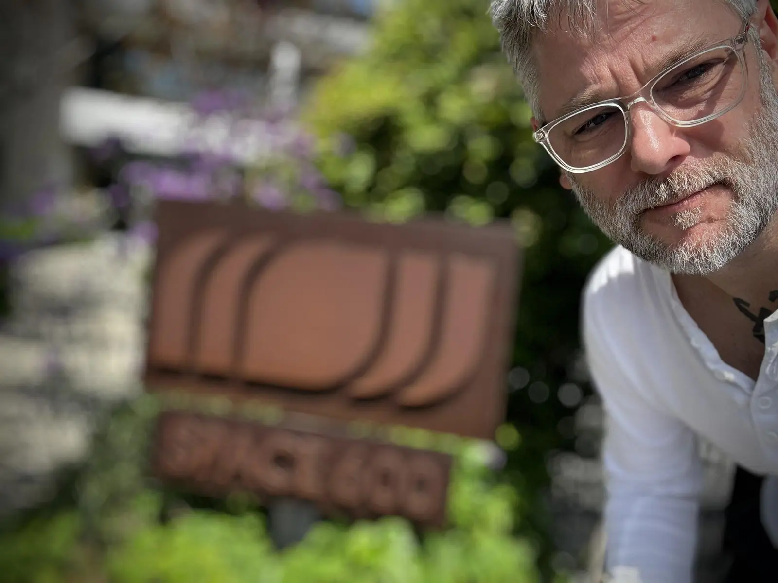 Portrait of Cory Barlog wearing glasses and a white t-shirt, with a blurred building sign in the background.