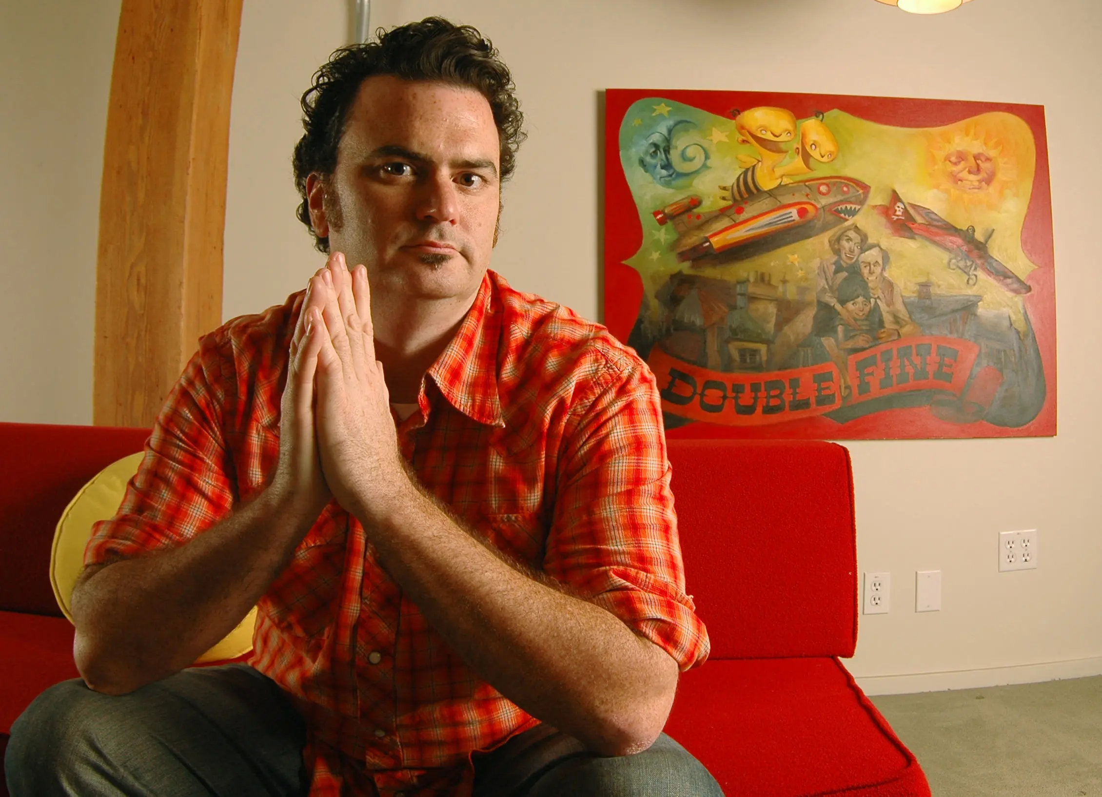 Tim Schafer in front of a Double Fine mural, posing with hands together.