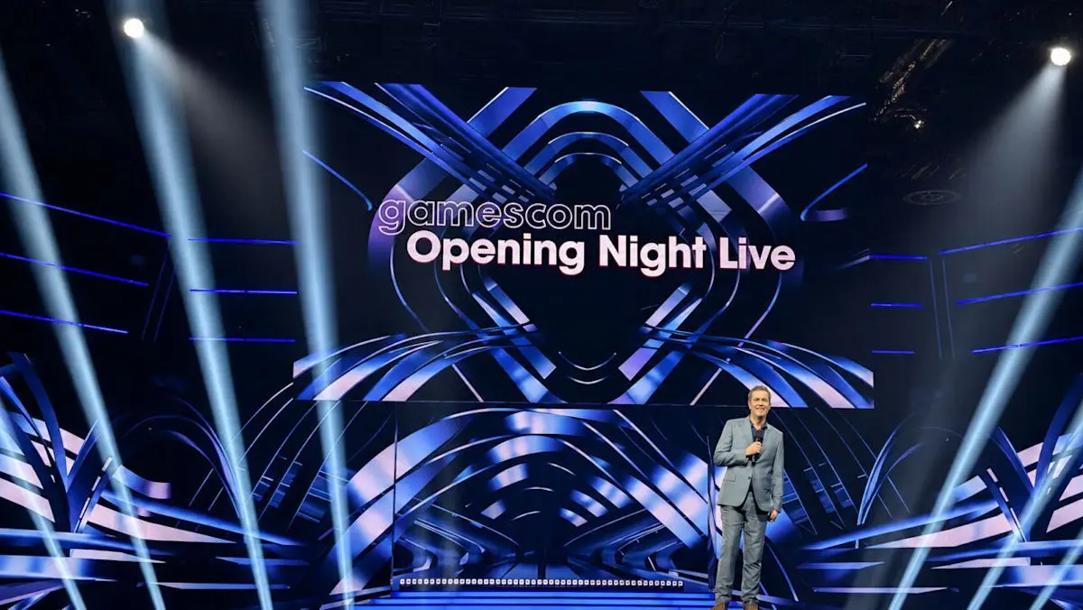 A presenter stands on the gamescom Opening Night Live stage with a large digital screen and blue spotlights.