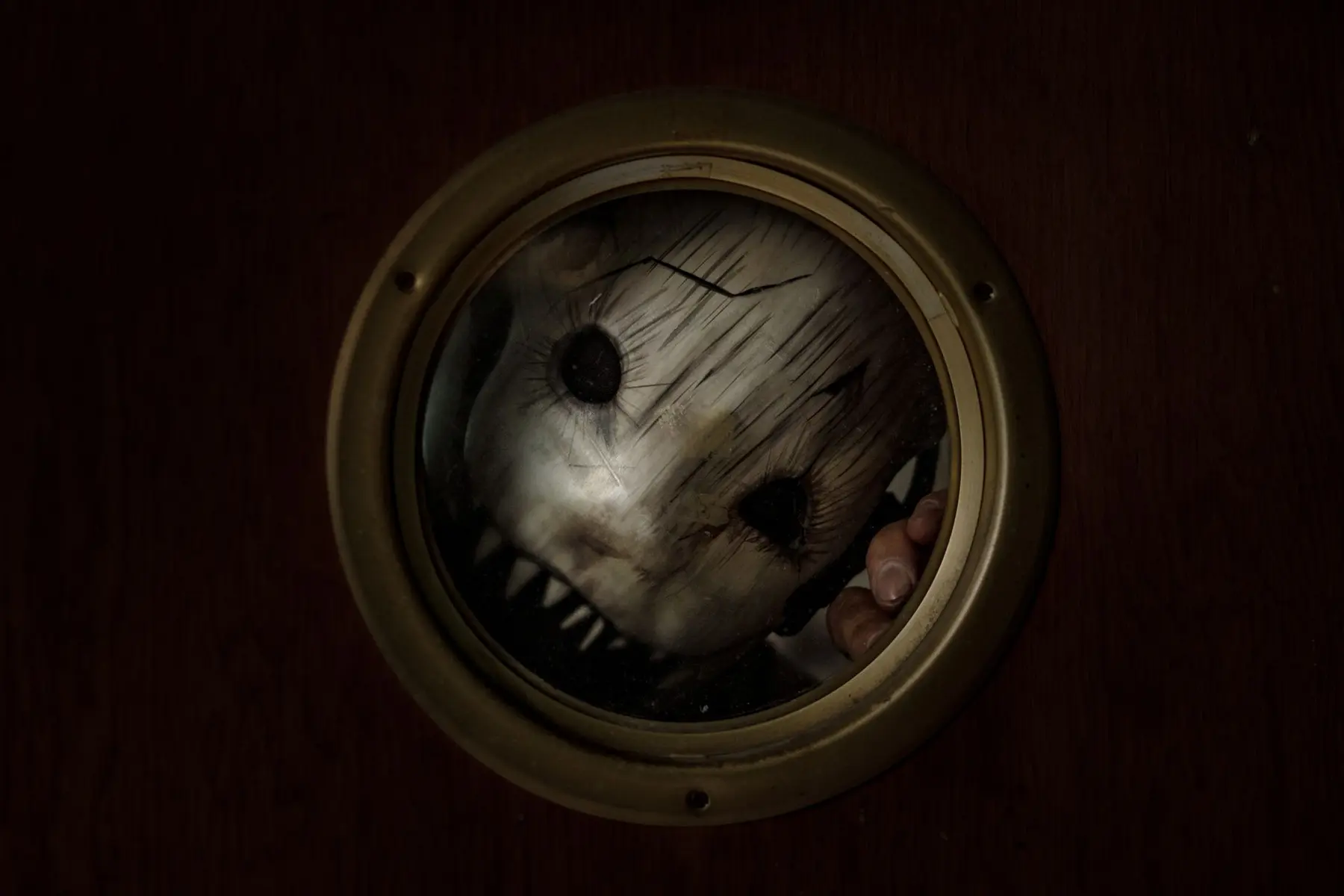 A cracked, pale horror mask with dark eye sockets peering through a circular brass porthole