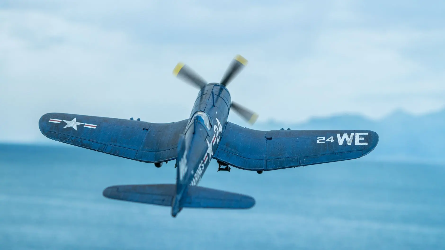 A blue F4U Corsair fighter plane in flight over a blue ocean with mountains in the distance